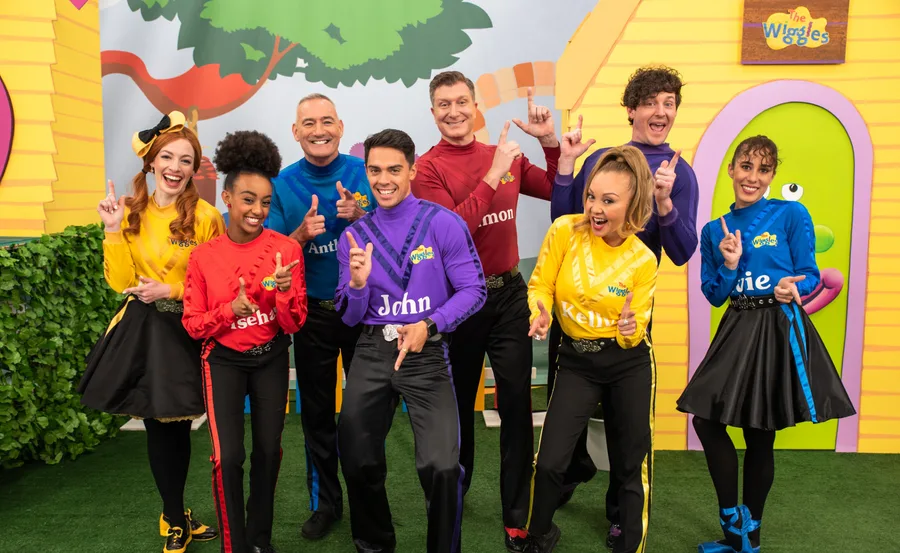 "The Wiggles group members dressed in colorful outfits with their names on shirts, smiling and posing enthusiastically."