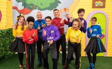 "The Wiggles group members dressed in colorful outfits with their names on shirts, smiling and posing enthusiastically."