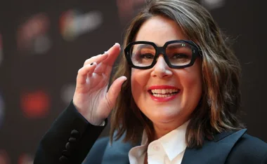 A smiling woman with glasses and a white shirt, holding her hand near her face on a dark background.