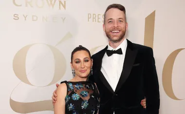 A smiling couple dressed formally at an event, with a beige backdrop featuring logos.