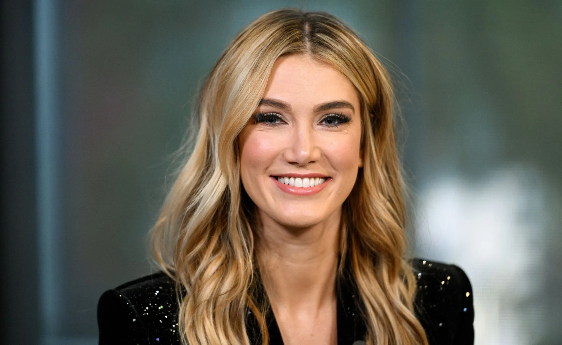 A person with long blonde hair and a sparkling jacket smiling at the camera.