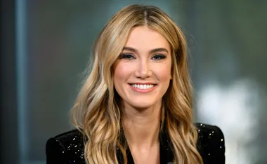 A person with long blonde hair and a sparkling jacket smiling at the camera.