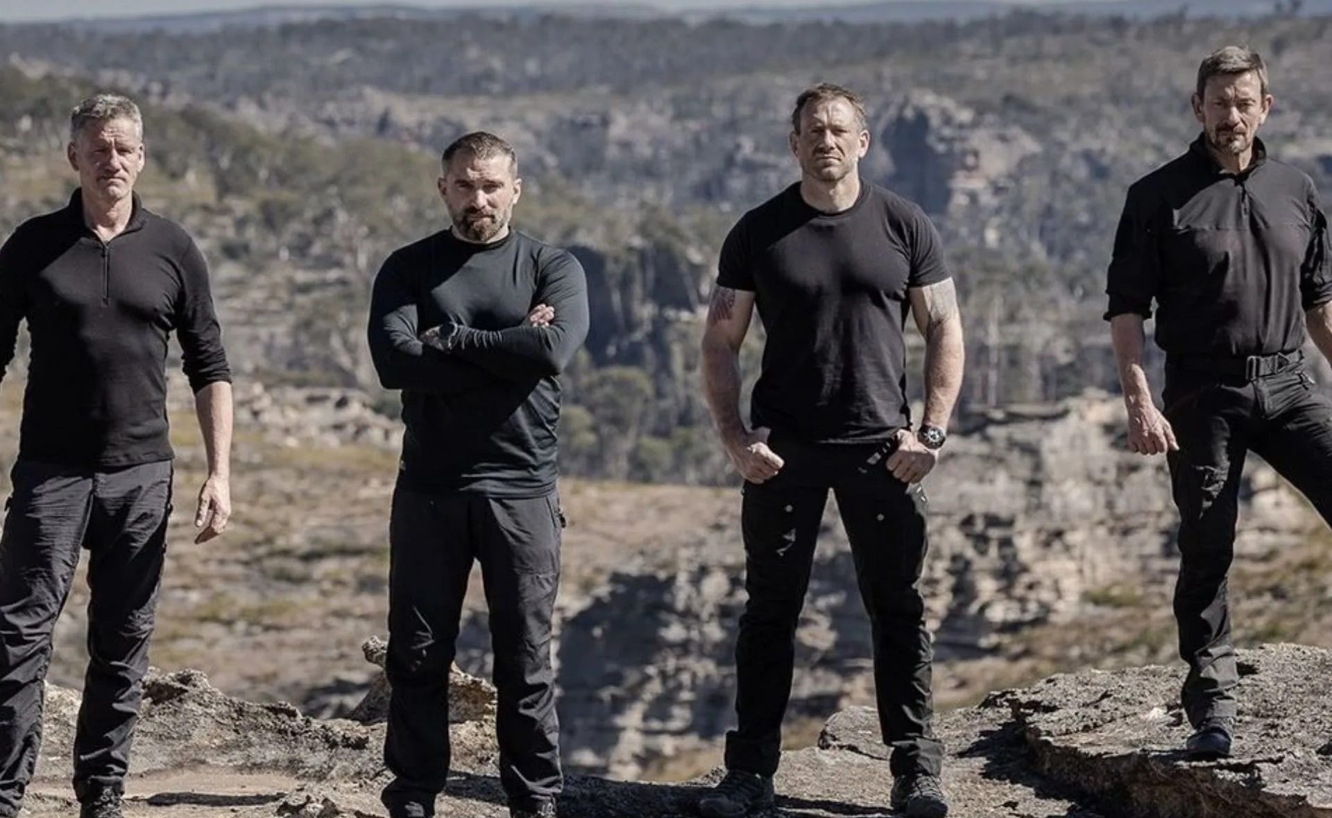 Four men in tactical outfits stand outdoors against a rugged, rocky landscape backdrop.