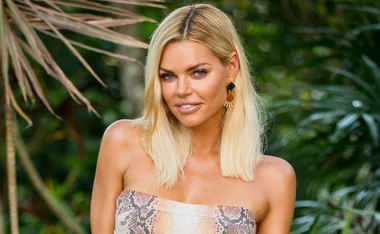Blonde woman with shoulder-length hair and patterned strapless top, standing in a lush outdoor setting.