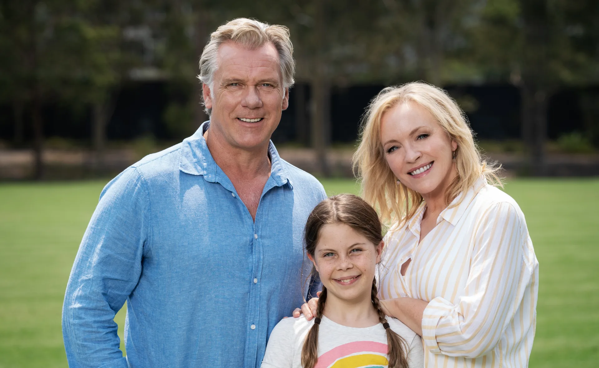 A smiling family of three, including a man, woman, and young girl, stand close together on a grassy field.