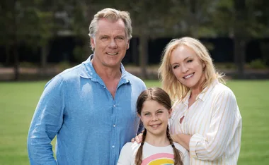 A smiling family of three, including a man, woman, and young girl, stand close together on a grassy field.