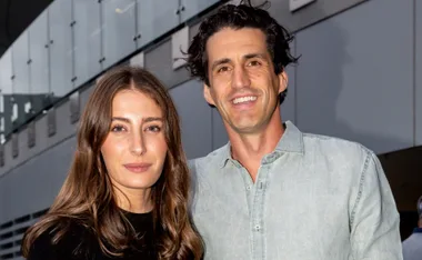 A smiling couple stands together outdoors, with a modern building in the background.