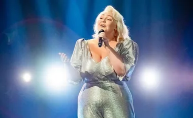 Singer in a silver outfit performs passionately on stage with bright lights in the background.