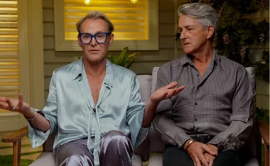 Two men sitting on a couch, one gesturing with hands, discussing something on a show set background.