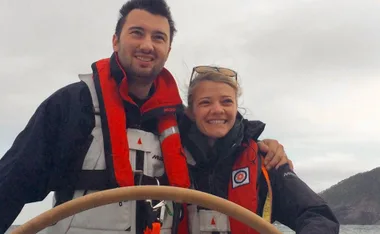 Two people smiling on a boat, both wearing life jackets, with cloudy skies and a coastline in the background.