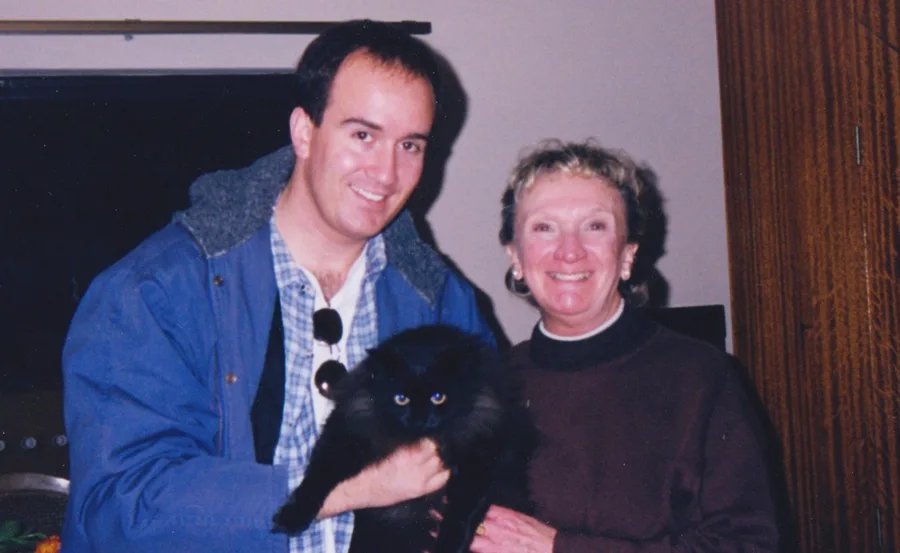Man and woman smiling, holding a black cat indoors.