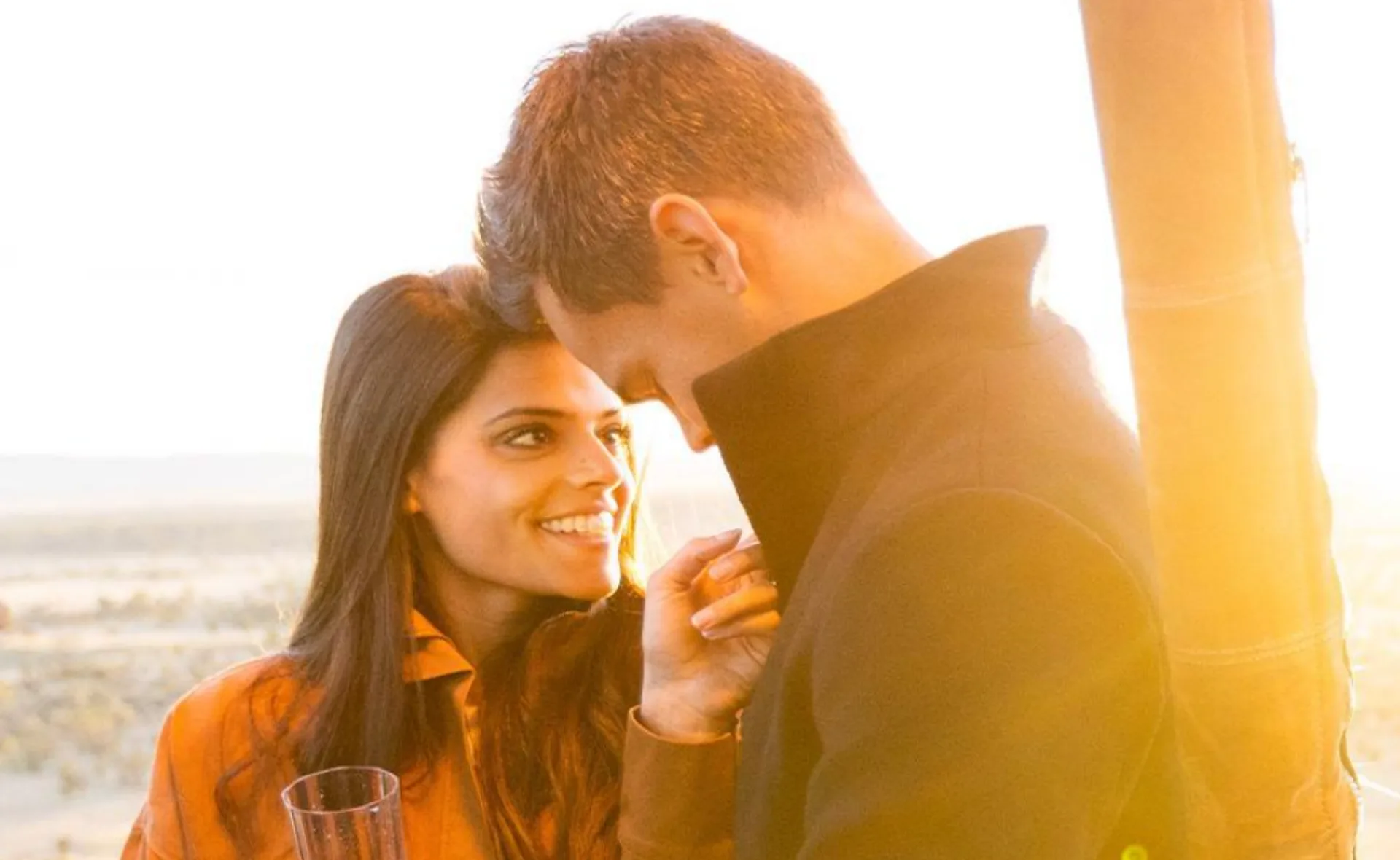 A woman smiles at a man during sunset, holding a champagne flute, with a warm, glowing background.