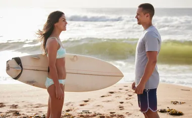 Two people on a beach, one holding a surfboard, facing each other with waves in the background.