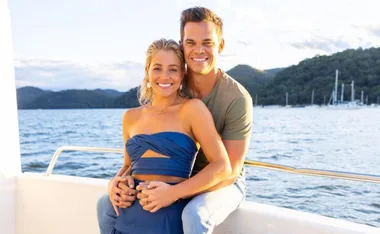 A couple smiling on a boat with a scenic view of water and hills in the background.