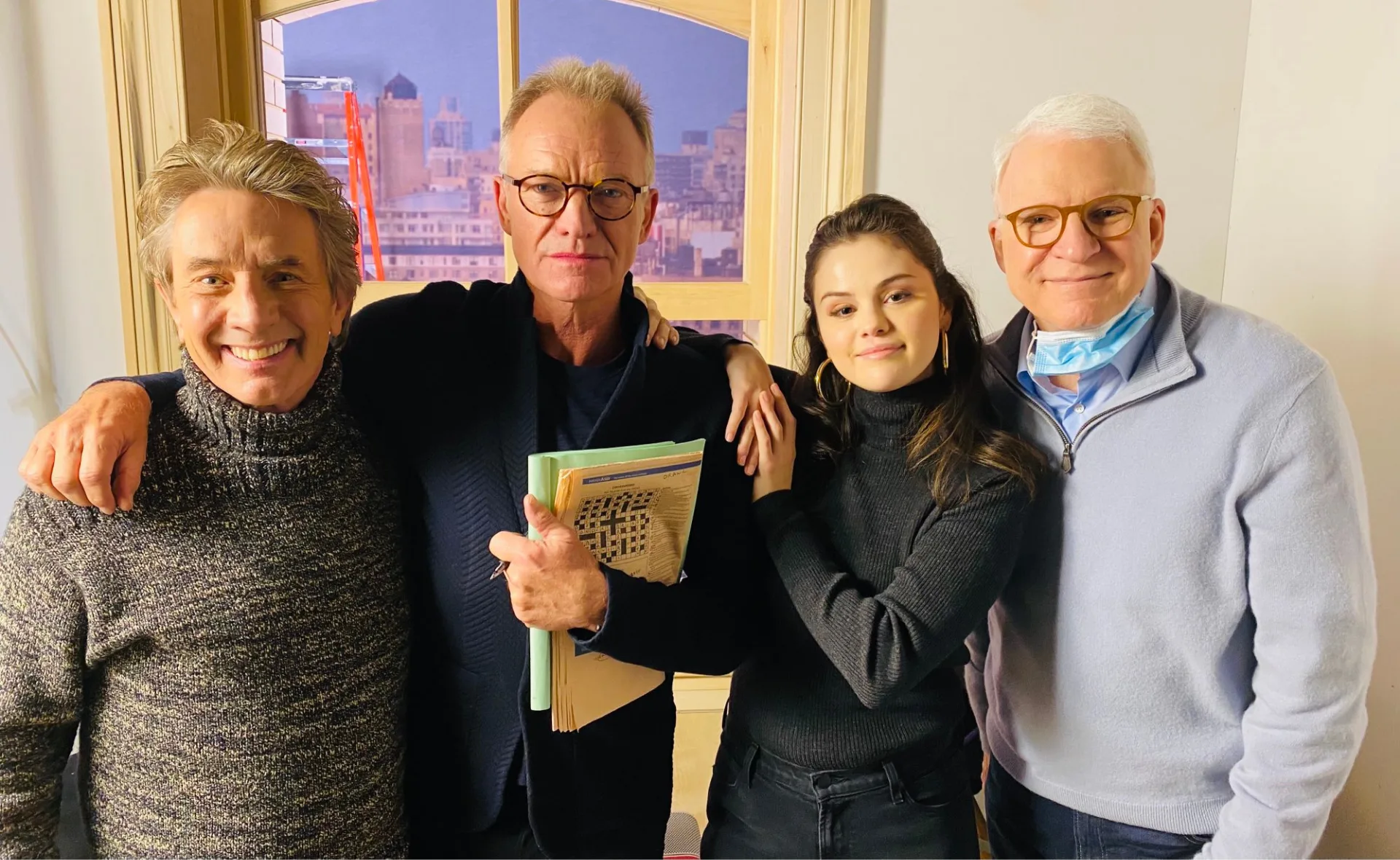 Four people smiling, one holds a crossword, standing in an apartment set from "Only Murders in the Building".