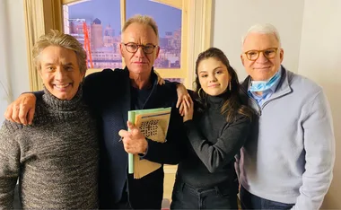 Four people smiling, one holds a crossword, standing in an apartment set from "Only Murders in the Building".
