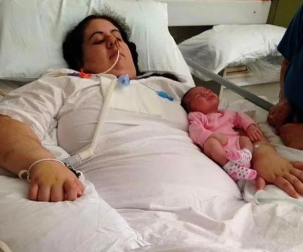 Woman in hospital bed with breathing tube, newborn baby in pink outfit resting beside her.