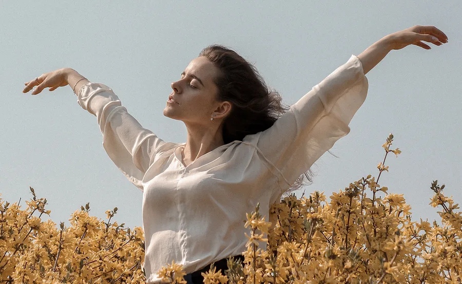 Woman with outstretched arms stands among blooming yellow flowers against a clear sky.