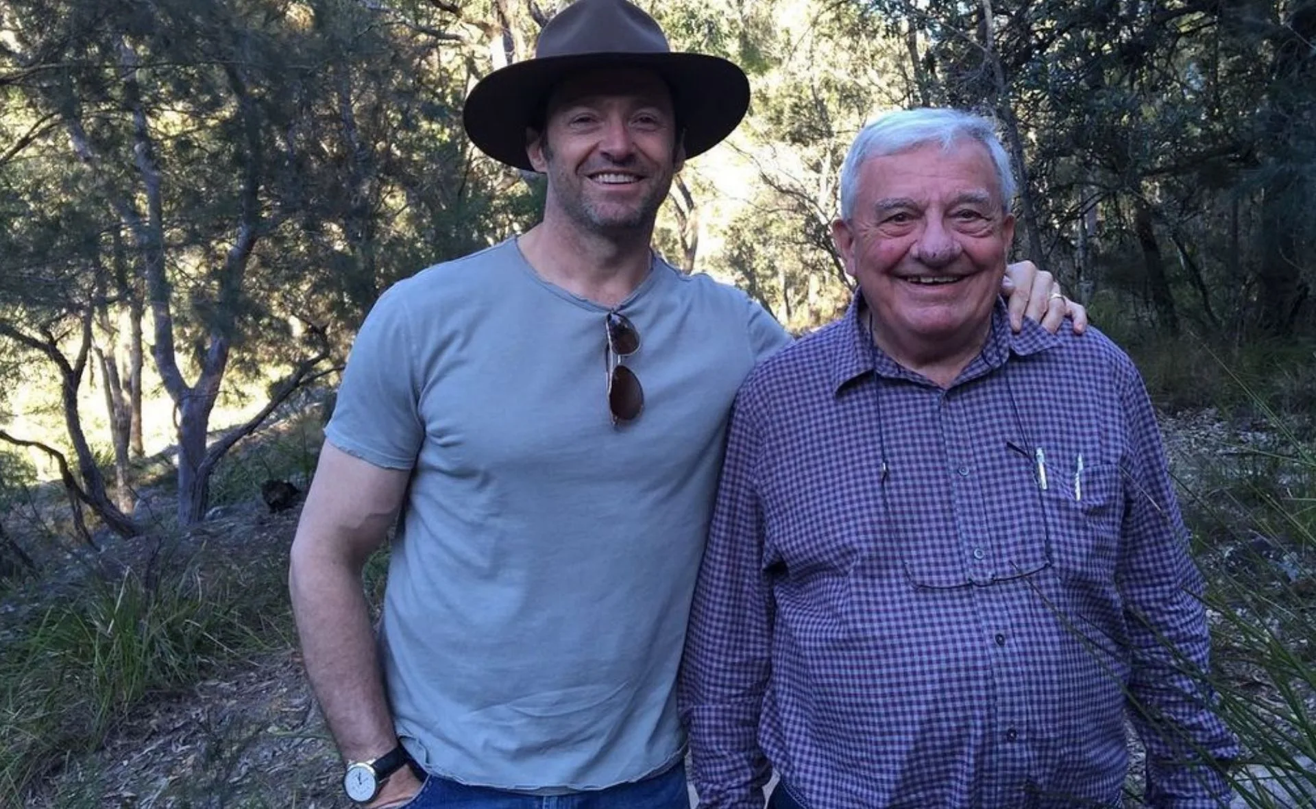 Two men smiling outdoors, one wearing a hat and sunglasses, standing close with trees in the background.