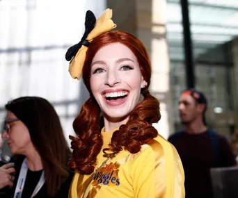 A woman in a yellow Wiggles costume with red hair and a yellow bow smiles brightly at an event.
