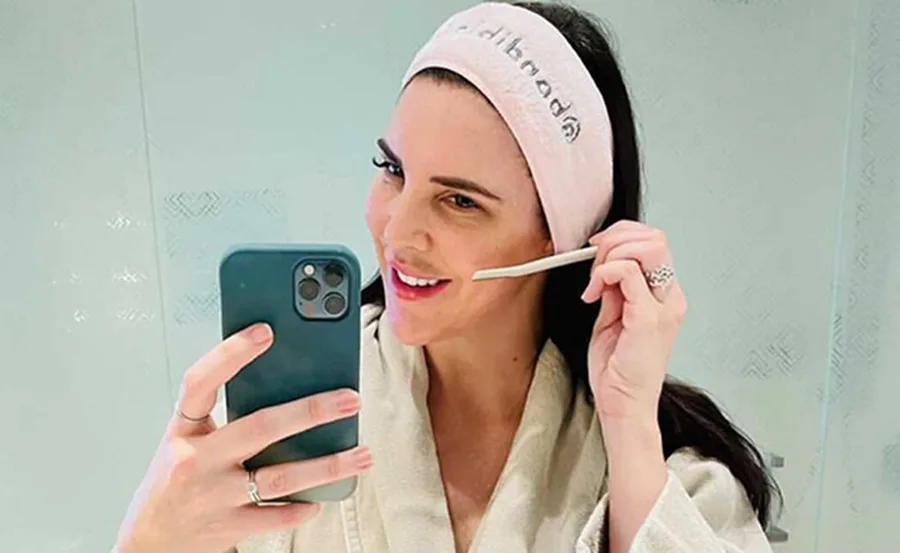 Woman dermaplaning her face in a bathroom mirror, holding a smartphone and wearing a pink headband.