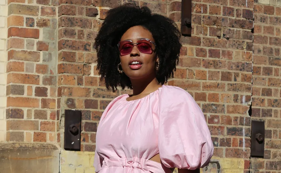 Person in pink dress and sunglasses against a brick wall.