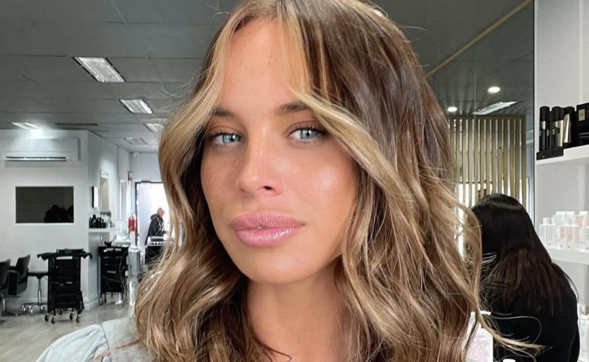 A woman with wavy hair in a salon, partially shown face, soft lighting, and modern decor in the background.