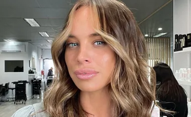 A woman with wavy hair in a salon, partially shown face, soft lighting, and modern decor in the background.