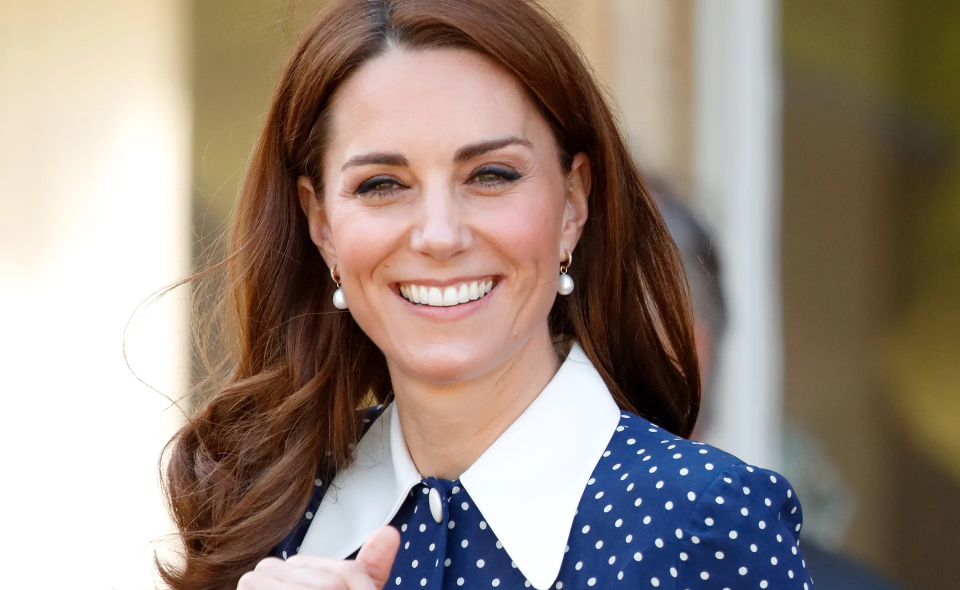 A woman with long brown hair, wearing a polka dot dress and pearl earrings, smiling outdoors.