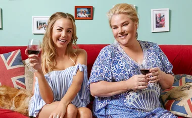 Two women sitting on a red couch, smiling with glasses of wine, with decorative pillows and framed photos in the background.