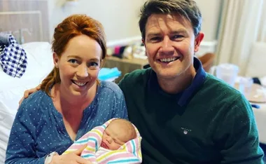 Smiling couple holding a newborn wrapped in a colorful blanket in a hospital room.