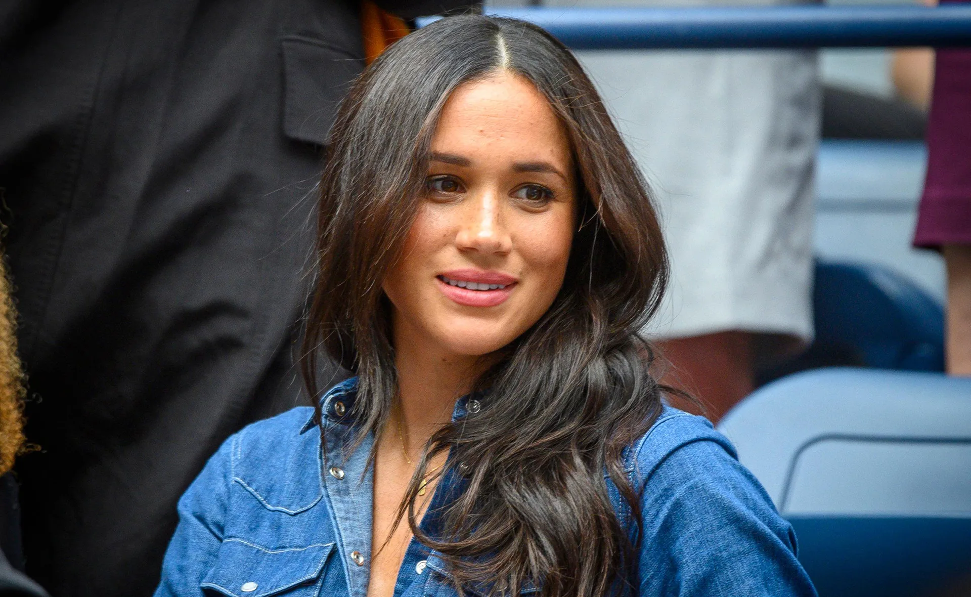 Woman with long hair in a blue denim shirt, smiling at an event with people in the background.
