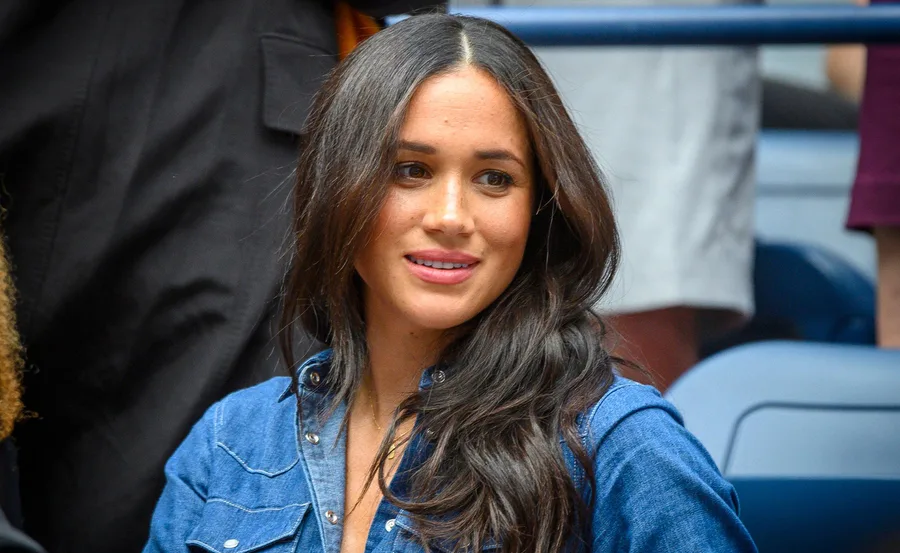 Woman with long hair in a blue denim shirt, smiling at an event with people in the background.