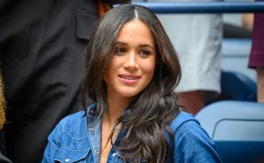 Woman with long hair in a blue denim shirt, smiling at an event with people in the background.