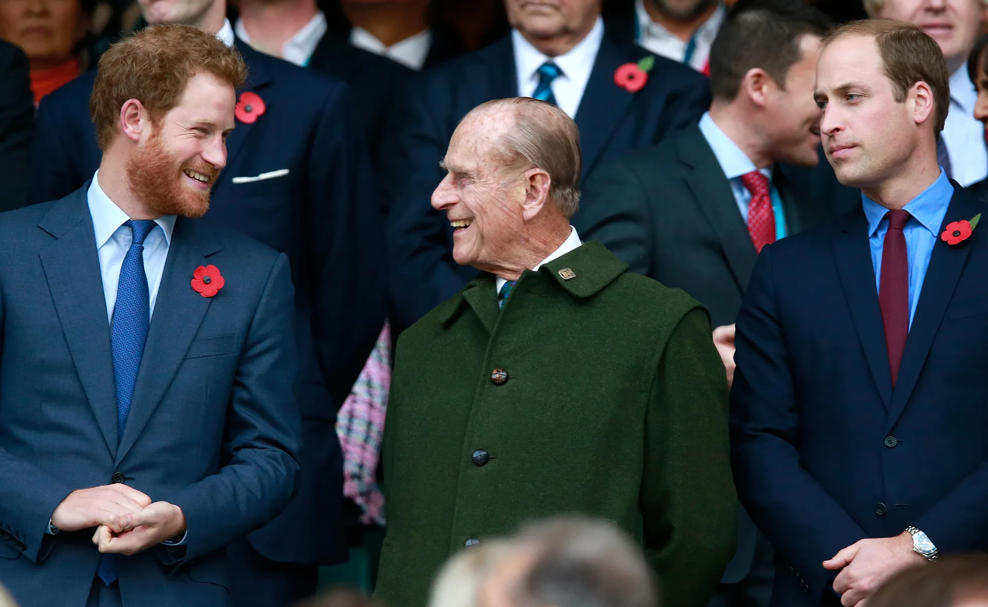 Three men in suits converse and smile at an event, each wearing a red poppy on their lapel.