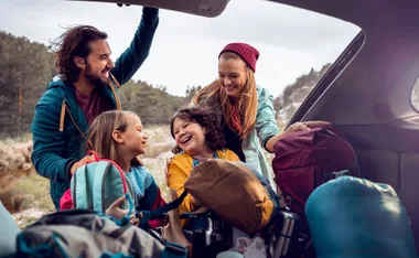 Family packing car trunk with backpacks for road trip in a forest setting, smiling and enjoying the moment.