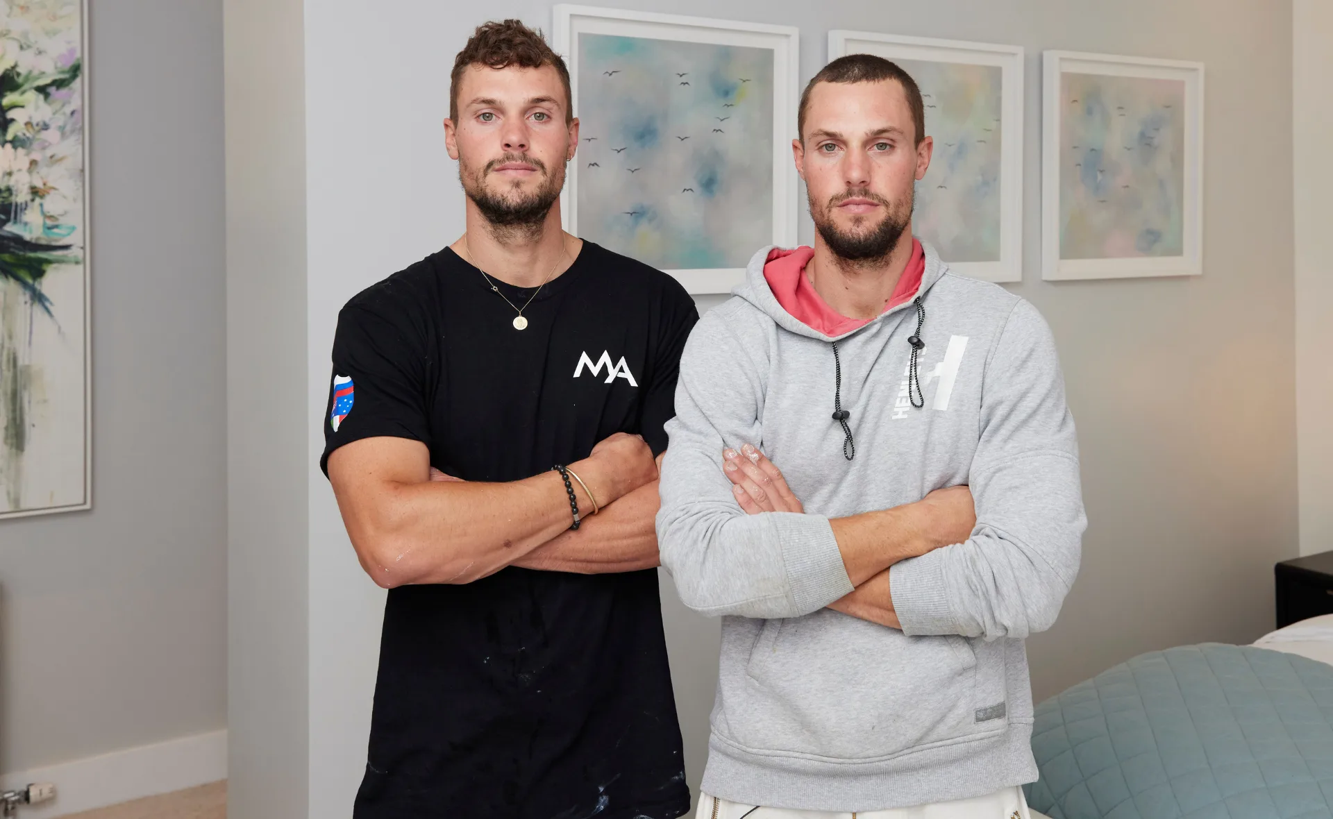 Two men standing side by side with arms crossed, in a room with abstract paintings on the wall.
