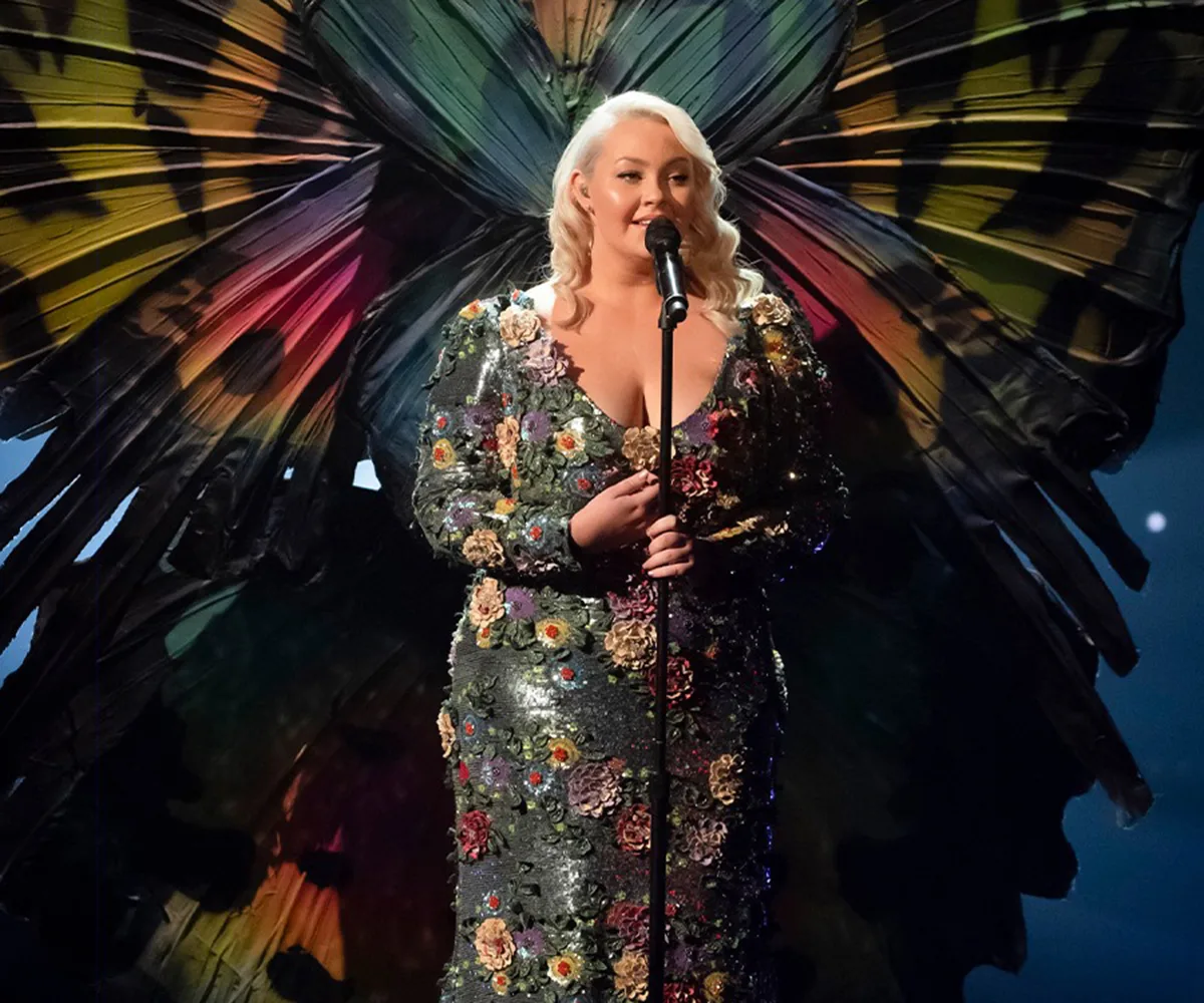 Singer in floral gown performs with butterfly wings backdrop on stage.
