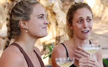 Two women from Australian Survivor 2021, one smiling and the other looking surprised, hold glasses outdoors.