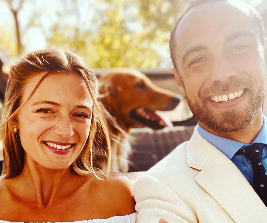 A smiling couple takes a selfie outdoors, with a dog in the background and sunlight streaming through.