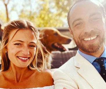 A smiling couple takes a selfie outdoors, with a dog in the background and sunlight streaming through.
