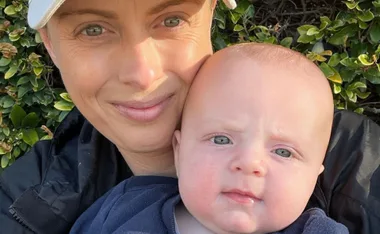 A person wearing a cap is holding a baby close, both looking at the camera with greenery in the background.