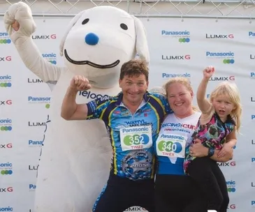 A family poses with a costumed character at a race event, wearing numbered bibs, smiling and celebrating.