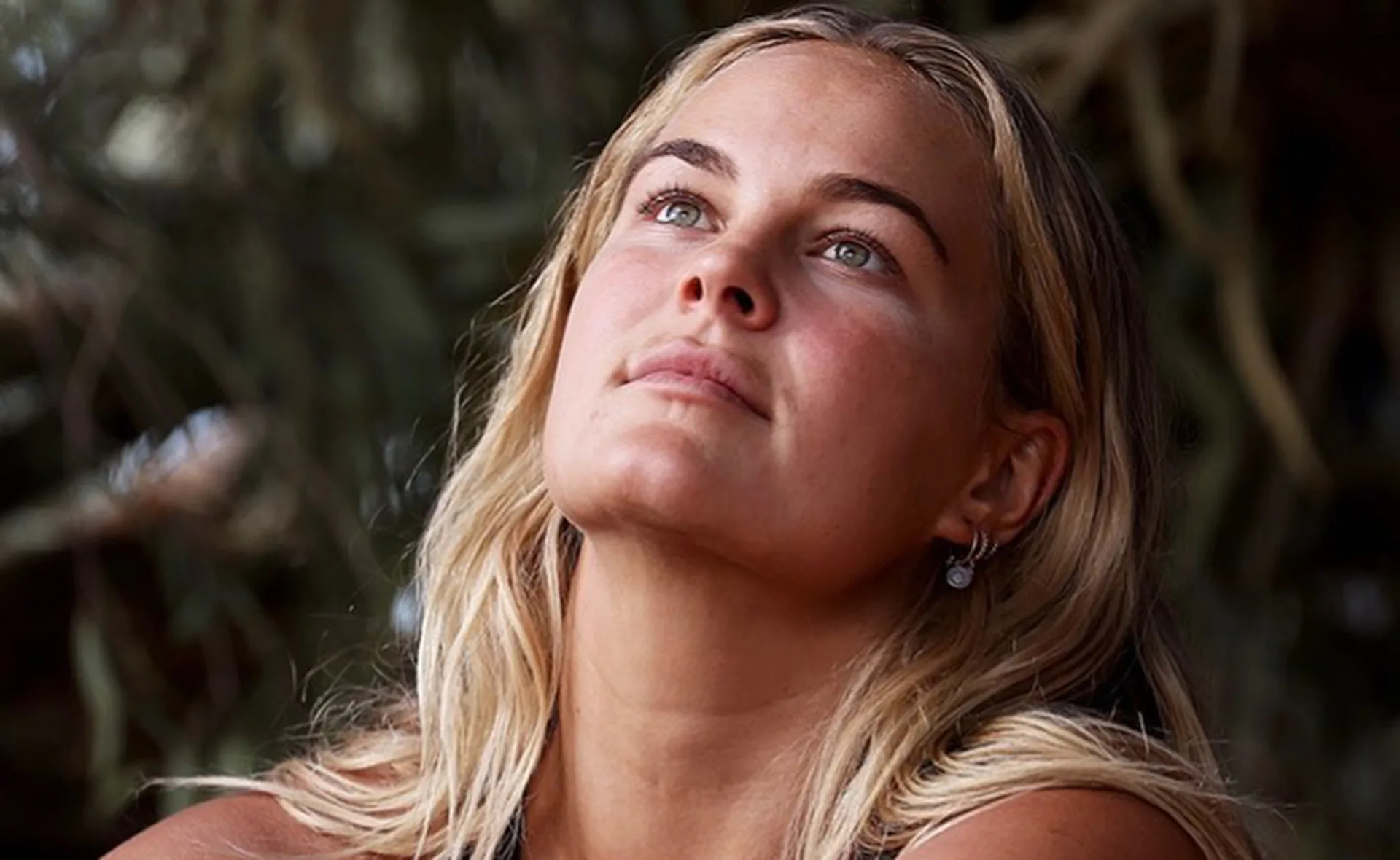 Woman with long blonde hair looks upward thoughtfully, surrounded by blurred foliage.