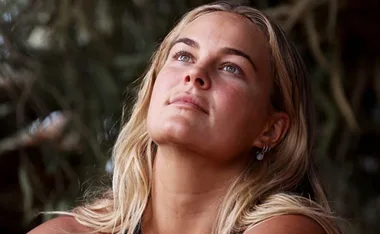 Woman with long blonde hair looks upward thoughtfully, surrounded by blurred foliage.