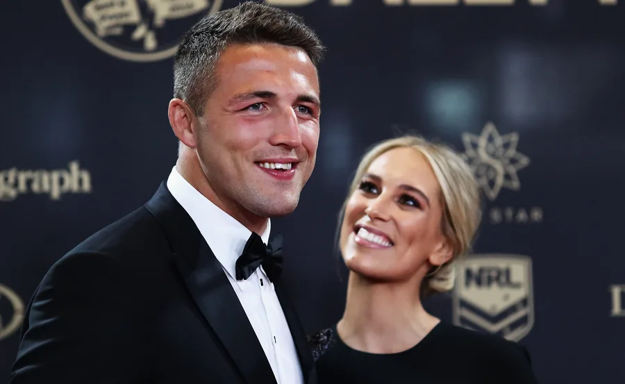 Two people smiling at an event, wearing formal attire in front of a branded backdrop.