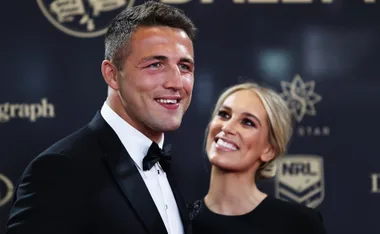 Two people smiling at an event, wearing formal attire in front of a branded backdrop.