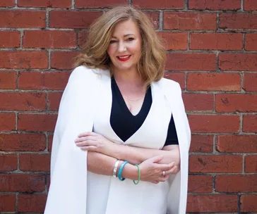 A woman with wavy hair in a white outfit, smiling in front of a red brick wall.