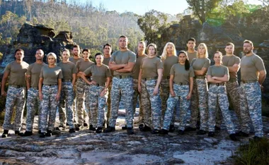 A group of 18 people in camo pants and brown shirts stand outdoors with rocky cliffs in the background.