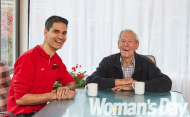 Two men smiling at a table with mugs, one in a red shirt, sitting indoors with a bright background.